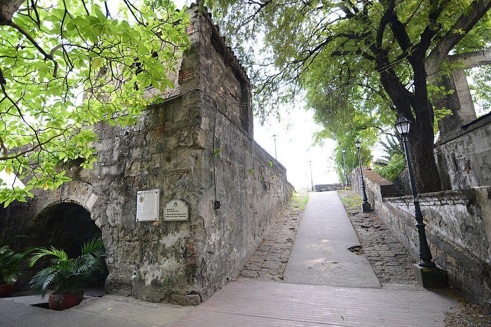 Historic stone walls of Intramuros in Manila, with a paved walkway and trees alongside the fortifications