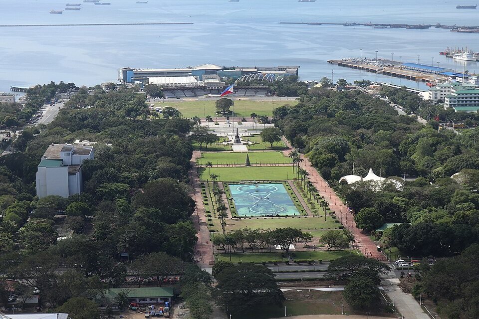Aerial view of Rizal Park in Manila on a clear day, showing its gardens, central fountain, tree-lined paths and Manila Bay