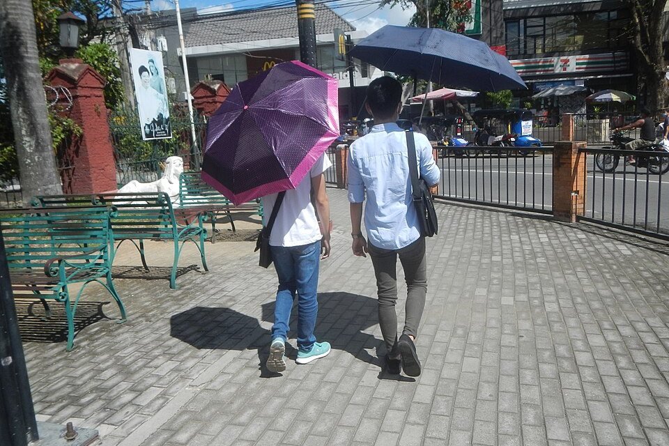 Two pedestrians carrying umbrellas for shade while walking along a sunlit pavement in Manila