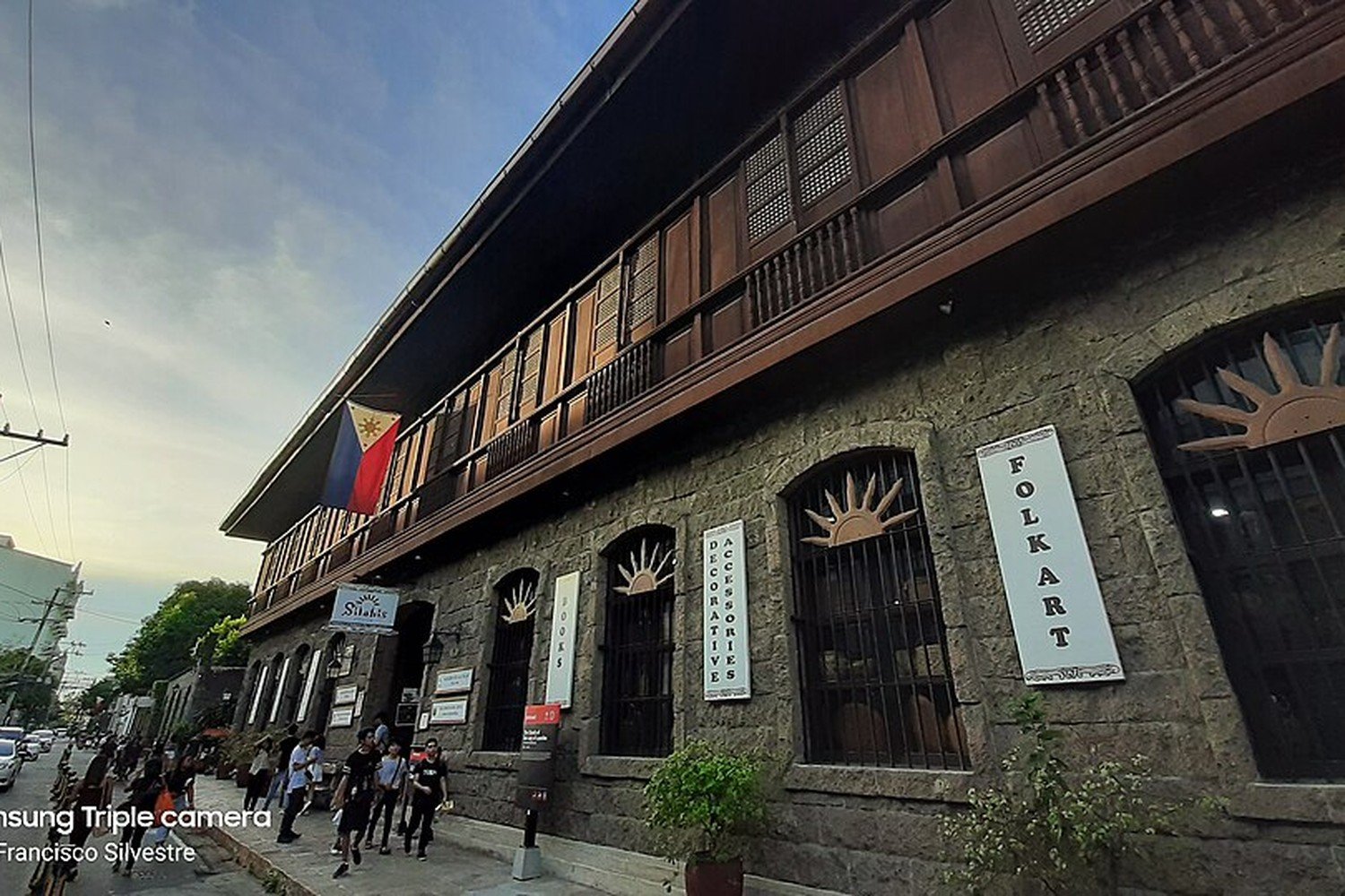 Historic stone building with wooden upper floors, pedestrians and a Philippine flag along a street in Intramuros, Manila