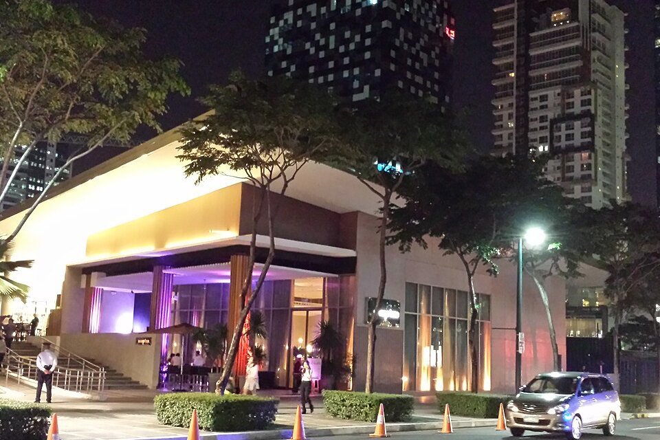 Illuminated building entrance at night with trees, a parked vehicle and high-rise buildings in the background