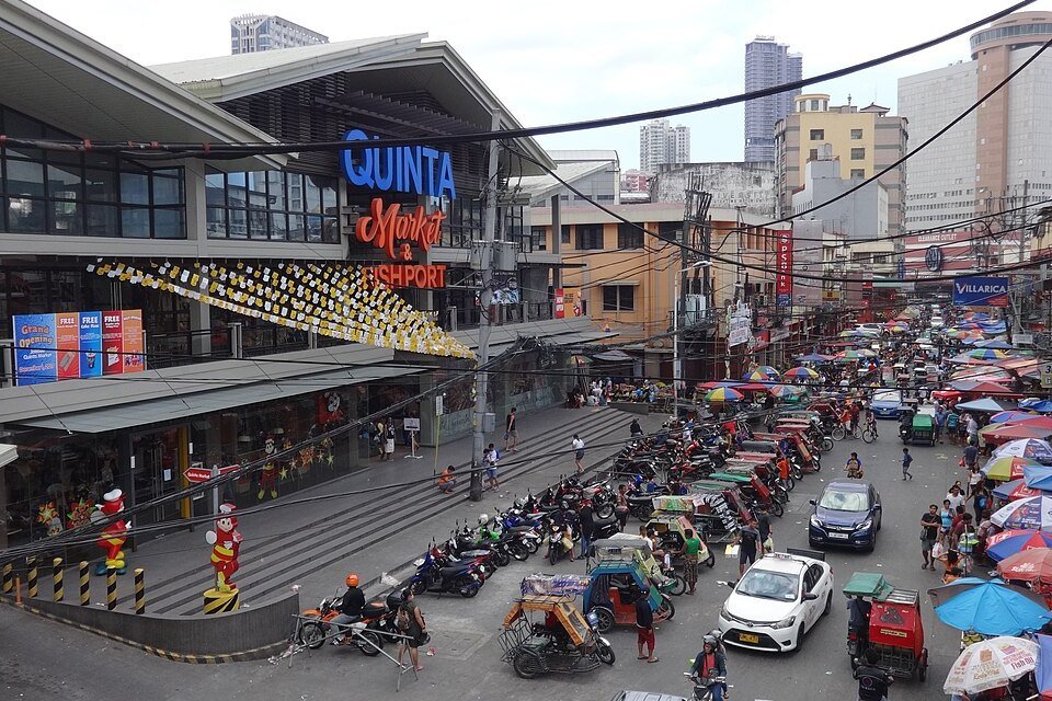 Busy street outside Quinta Market in Quiapo, Manila, with market stalls, colorful umbrellas, pedestrians and vehicles
