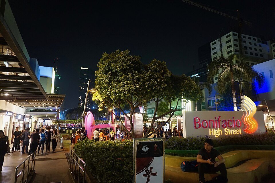 Bonifacio High Street in BGC at night, with illuminated shops, pedestrians, trees and the shopping district sign