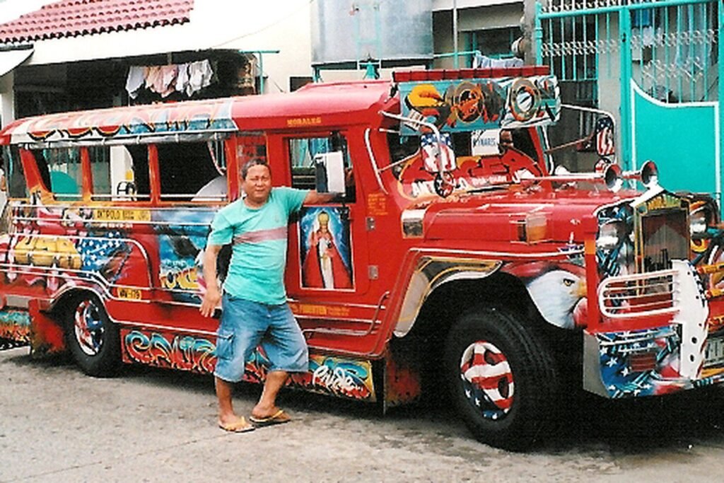 Man standing beside a brightly decorated red Filipino jeepney parked on a street