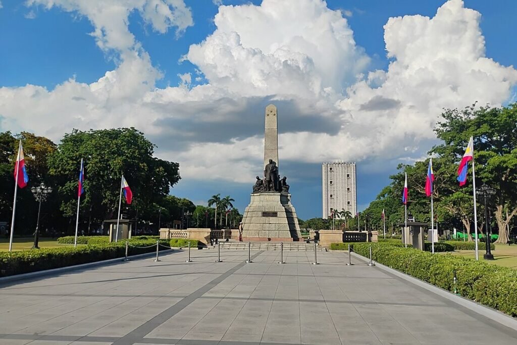 Wide paved walkway leading to the Rizal Monument, lined with Philippine flags and trees under a cloudy blue sky