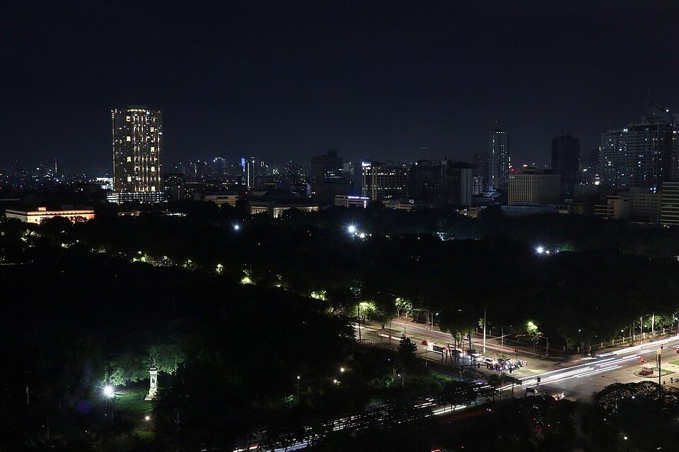 Night view across Rizal Park in Manila, with illuminated roads, trees and high-rise buildings in the distance